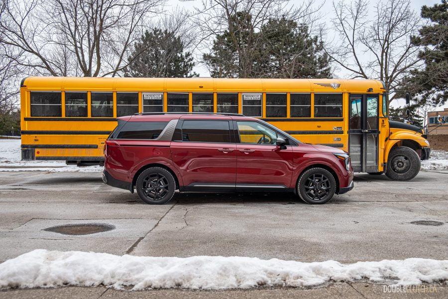 Side profile view of a red 2025 Kia Carnival Hybrid parked beside a school bus
