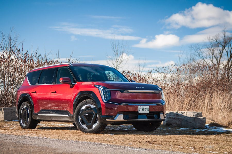 Front quarter view of a red 2025 Kia EV9 on a sunny day, with trees and shrubs in the background
