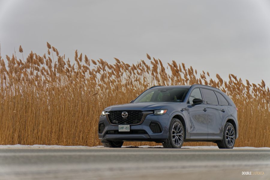 Front quarter view of a grey 2025 Mazda CX-70 GT PHEV on a cloudy day with trees in the background and snow on the ground
