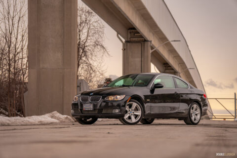 Front quarter view of a black 2008 BMW 335i Coupe on a cloudy day with an overpass in the background