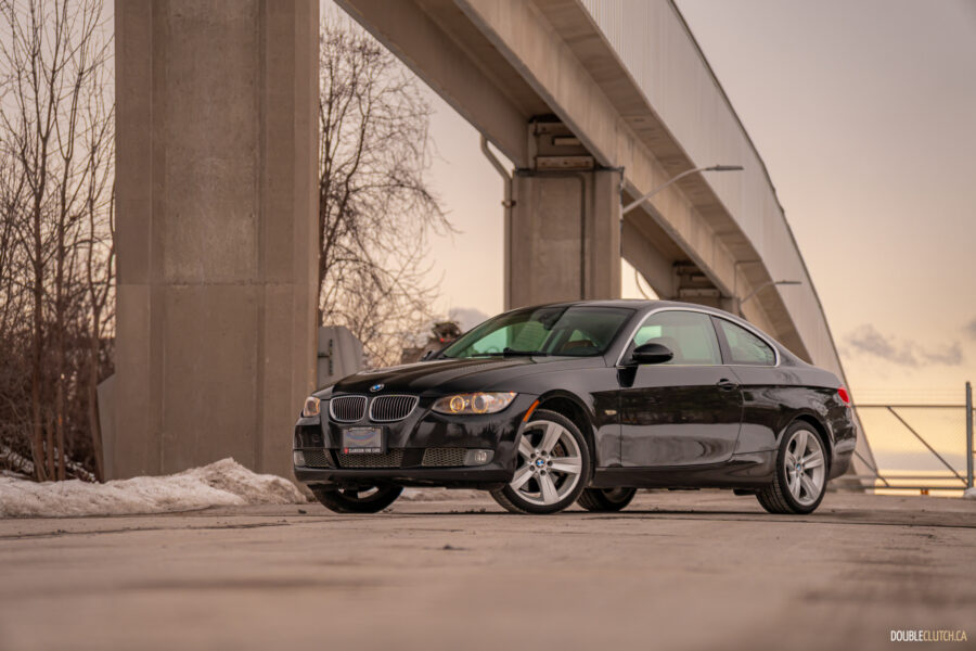 Front quarter view of a black 2008 BMW 335i Coupe on a cloudy day with an overpass in the background