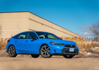 Front quarter view of a blue 2025 Honda Civic Hybrid Hatchback on a sunny day, with trees and a building in the background