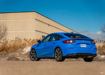 Rear quarter view of a blue 2025 Honda Civic Hybrid Hatchback on a sunny day, with trees and a building in the background