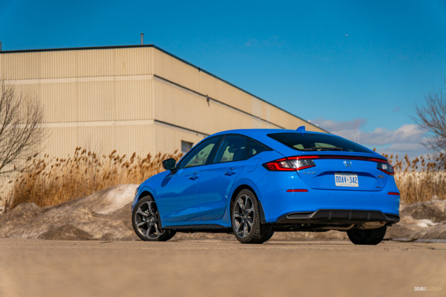 Rear quarter view of a blue 2025 Honda Civic Hybrid Hatchback on a sunny day, with trees and a building in the background