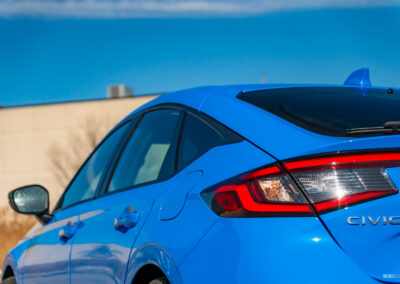 Rear quarter close-up view of a blue 2025 Honda Civic Hybrid Hatchback on a sunny day, with trees and a building in the background