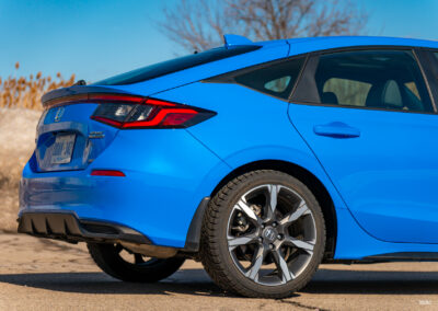 Rear quarter view of a blue 2025 Honda Civic Hybrid Hatchback on a sunny day, with trees and a building in the background