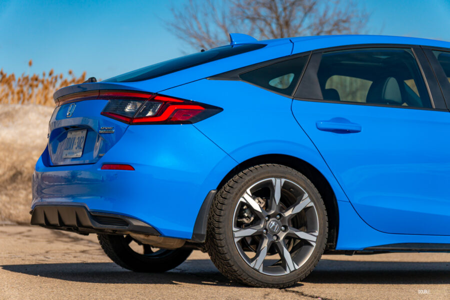 Rear quarter view of a blue 2025 Honda Civic Hybrid Hatchback on a sunny day, with trees and a building in the background