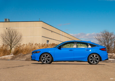 Side profile view of a blue 2025 Honda Civic Hybrid Hatchback on a sunny day, with trees and a building in the background