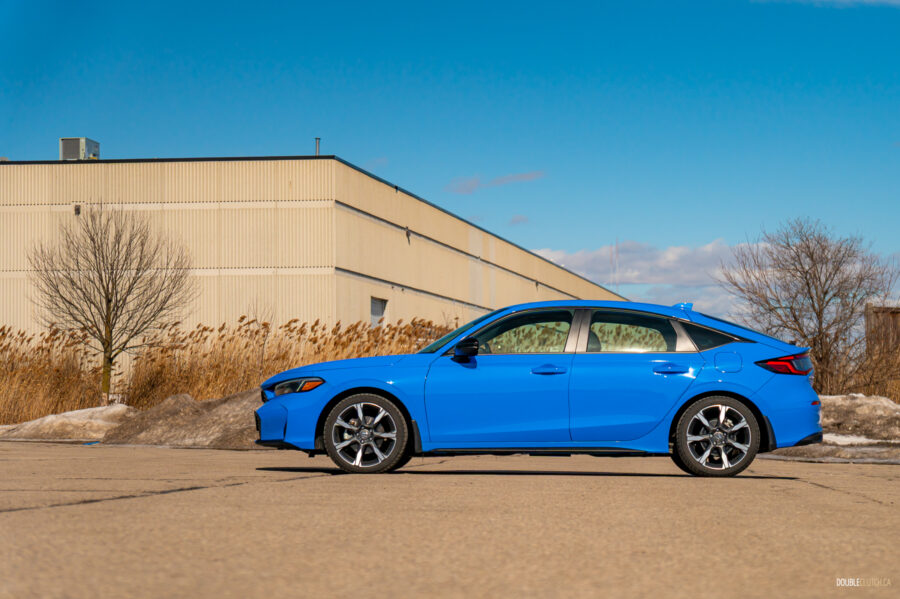 Side profile view of a blue 2025 Honda Civic Hybrid Hatchback on a sunny day, with trees and a building in the background