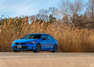 Front quarter view of a blue 2025 Honda Civic Hybrid Hatchback on a sunny day, with trees and shrubs in the background