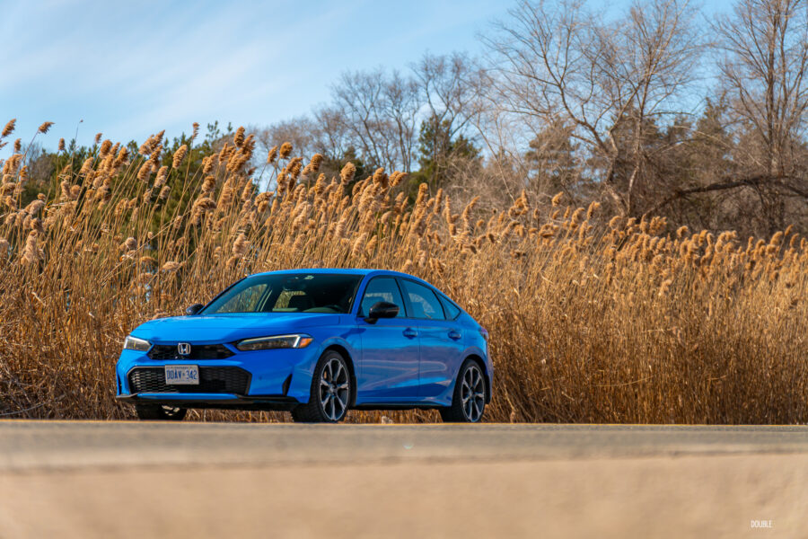 Front quarter view of a blue 2025 Honda Civic Hybrid Hatchback on a sunny day, with trees and shrubs in the background