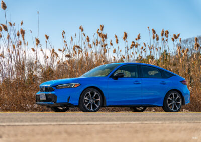 Front quarter view of a blue 2025 Honda Civic Hybrid Hatchback on a sunny day, with trees and shrubs in the background