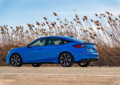 Rear quarter view of a blue 2025 Honda Civic Hybrid Hatchback on a sunny day, with trees and shrubs in the background
