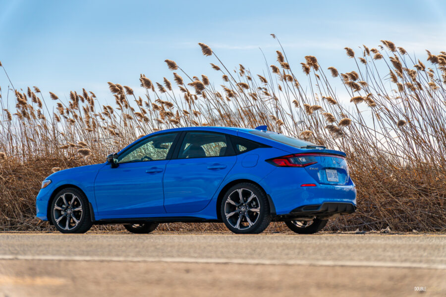 Rear quarter view of a blue 2025 Honda Civic Hybrid Hatchback on a sunny day, with trees and shrubs in the background