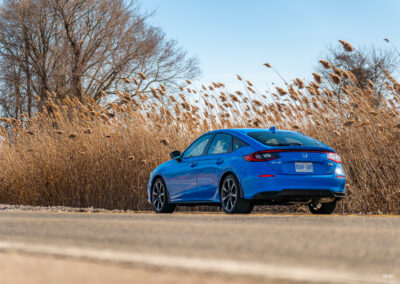 Rear quarter view of a blue 2025 Honda Civic Hybrid Hatchback on a sunny day, with trees and shrubs in the background