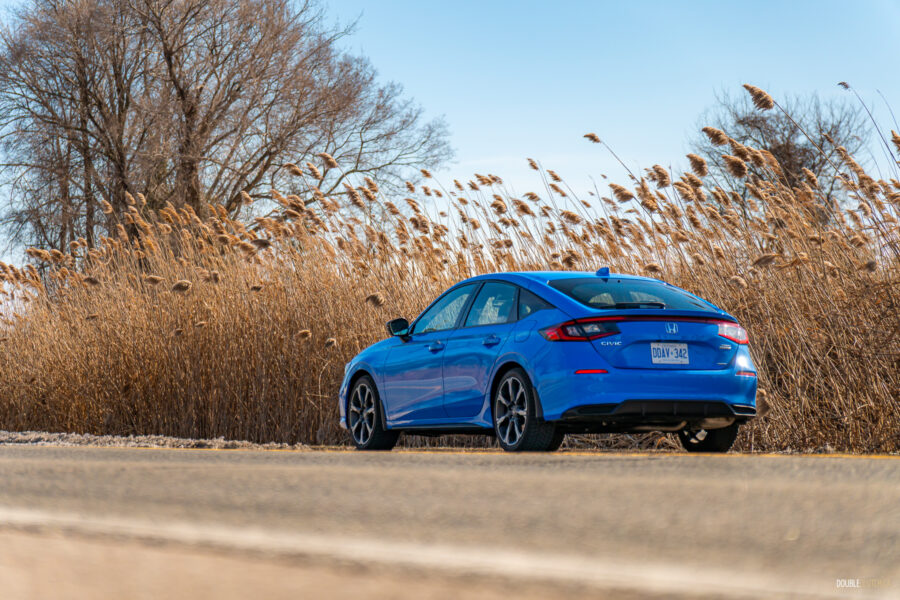 Rear quarter view of a blue 2025 Honda Civic Hybrid Hatchback on a sunny day, with trees and shrubs in the background