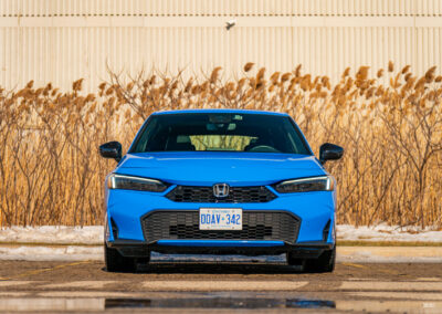 Front view of a blue 2025 Honda Civic Hybrid Hatchback on a sunny day, with trees and shrubs in the background
