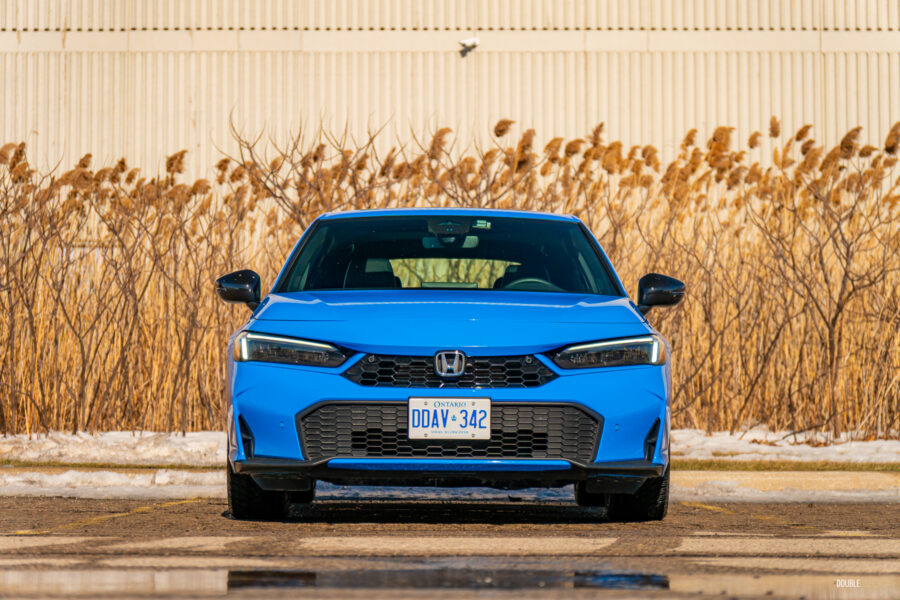 Front view of a blue 2025 Honda Civic Hybrid Hatchback on a sunny day, with trees and shrubs in the background