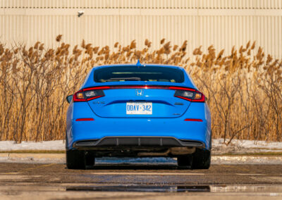 Rear view of a blue 2025 Honda Civic Hybrid Hatchback on a sunny day, with shrubs and a building in the background