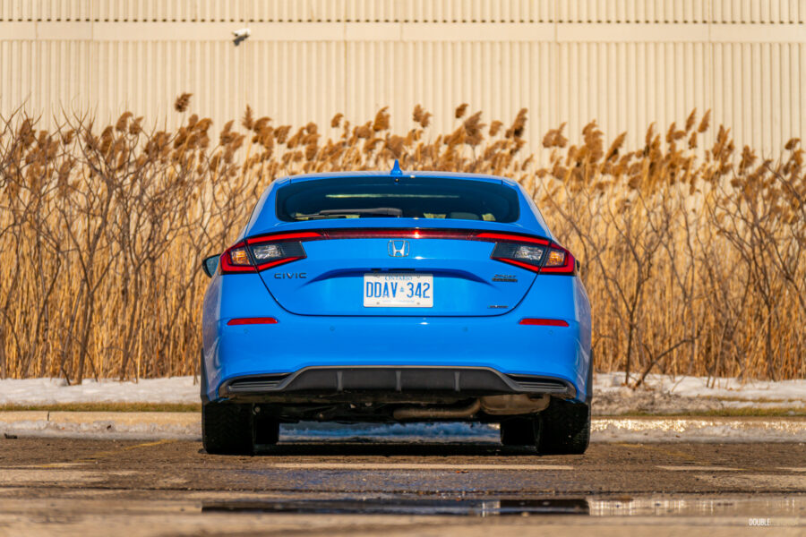 Rear view of a blue 2025 Honda Civic Hybrid Hatchback on a sunny day, with shrubs and a building in the background