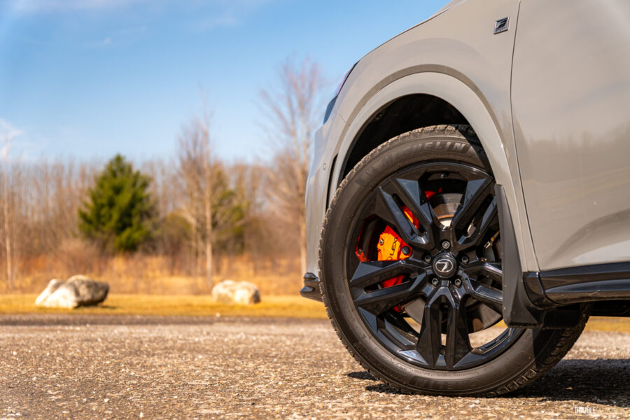 Close-up view of the gloss black front wheel and orange brake calipers on a 2025 Lexus RX 500h Black Line Special Edition on a sunny day with trees in the background