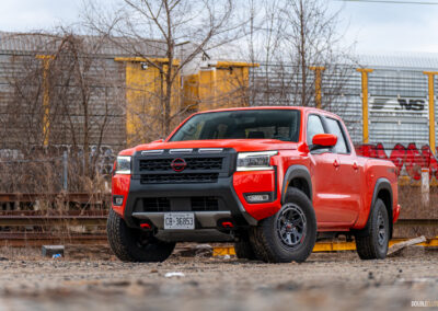 Front quarter view of an orange 2025 Nissan Frontier PRO-4X Luxury on a cloudy day, with trees and a train in the background