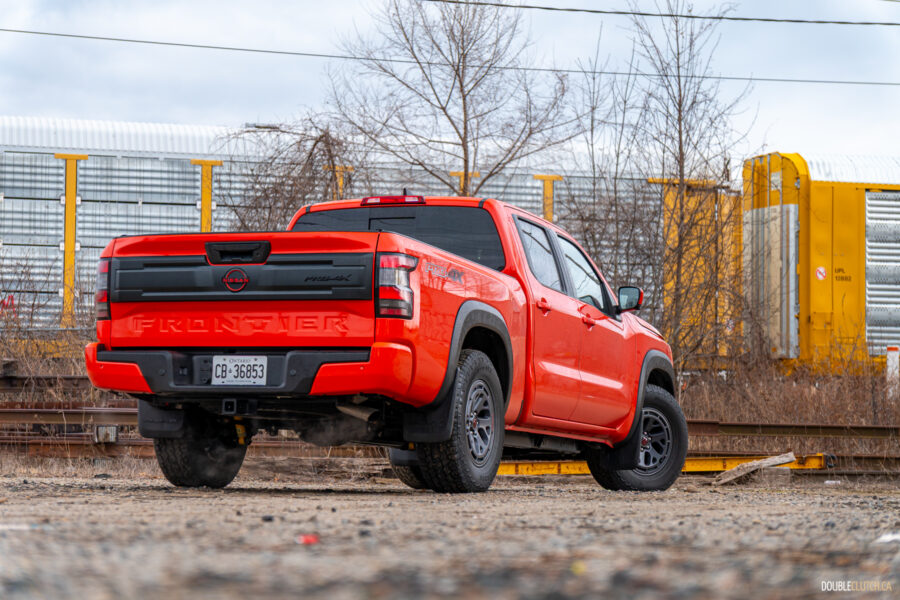 Rear quarter view of an orange 2025 Nissan Frontier PRO-4X Luxury on a cloudy day, with trees and a train in the background