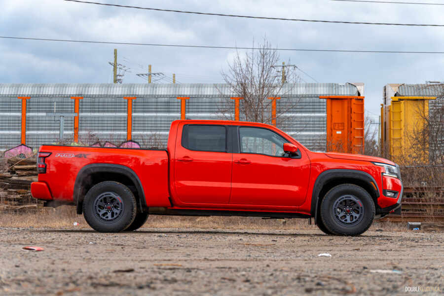Side profile view of an orange 2025 Nissan Frontier PRO-4X Luxury on a cloudy day, with trees and a train in the background
