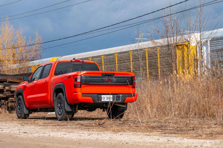 Rear quarter view of an orange 2025 Nissan Frontier PRO-4X Luxury on a cloudy day, with trees and a train in the background