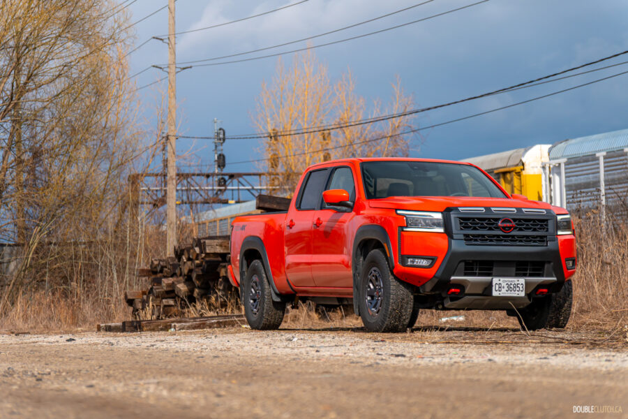 Front quarter view of an orange 2025 Nissan Frontier PRO-4X Luxury on a cloudy day, with trees and a train in the background