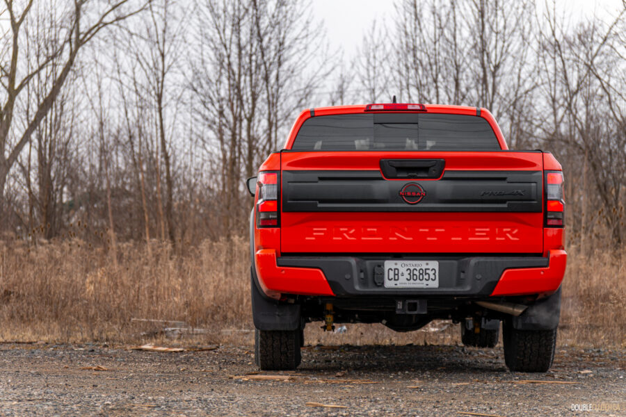 Rear view of an orange 2025 Nissan Frontier PRO-4X Luxury on a cloudy day, with trees and a train in the background