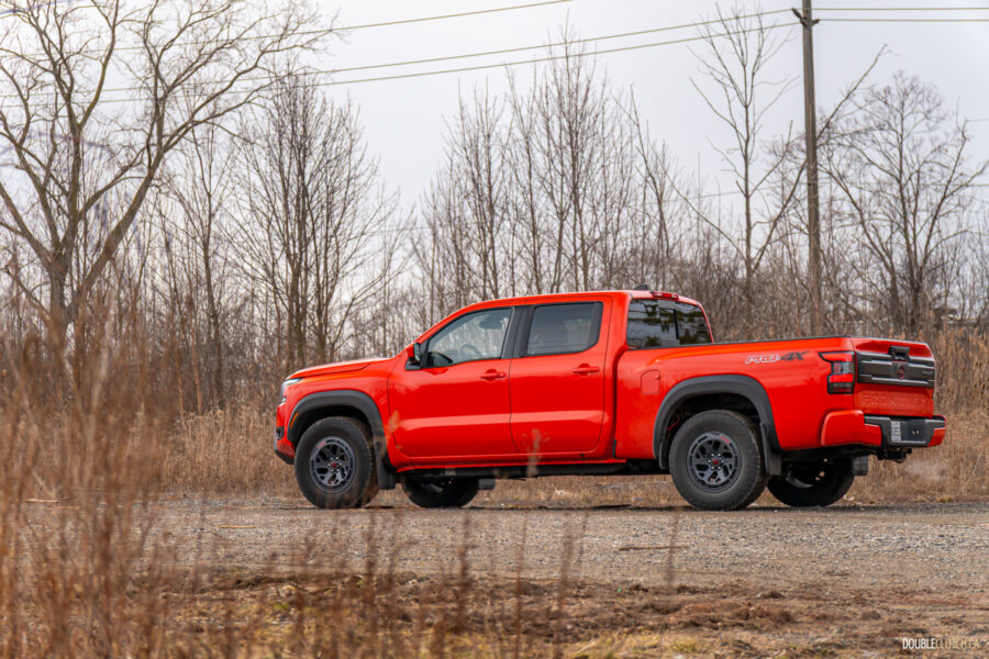 Rear quarter view of an orange 2025 Nissan Frontier PRO-4X Luxury on a cloudy day, with trees in the background