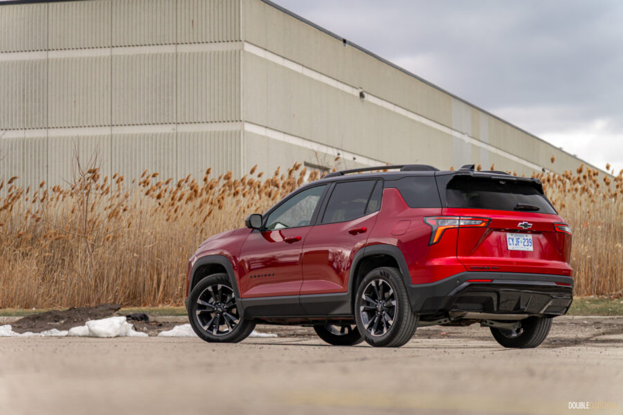 Rear quarter view of a red 2025 Chevrolet Equinox RS with a building and shrubs in the background