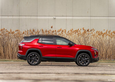 Side profile view of a red 2025 Chevrolet Equinox RS with a building and shrubs in the background