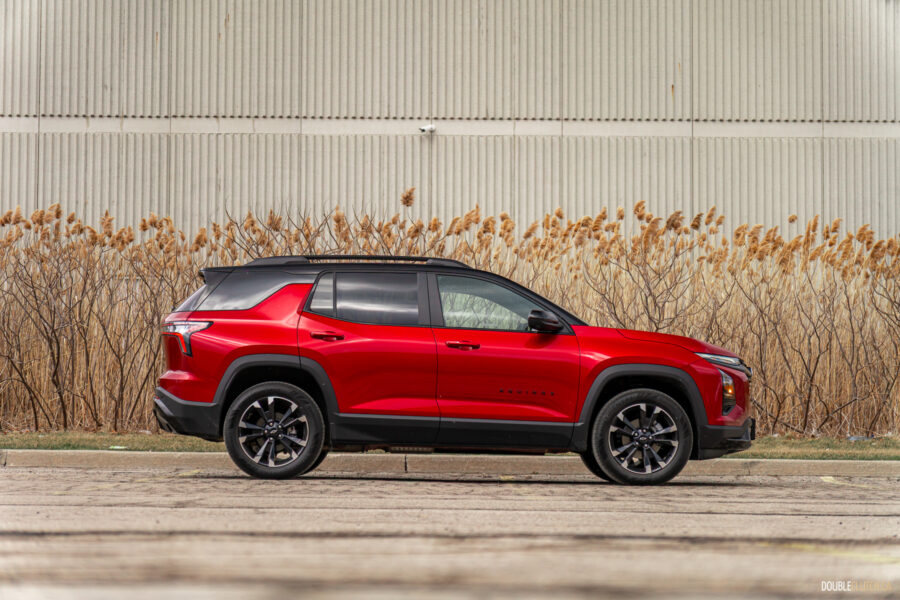 Side profile view of a red 2025 Chevrolet Equinox RS with a building and shrubs in the background