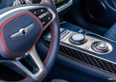 Close-up showing the steering wheel, carbon fibre-like trim, infotainment knob, and shifter dial arrangement in a 2025 Genesis GV70 3.5T Sport Plus
