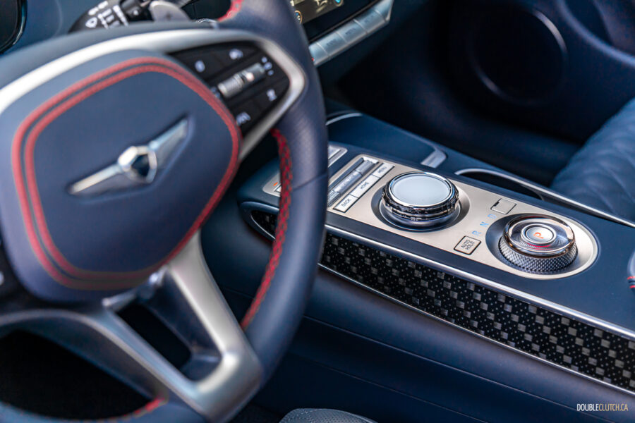 Close-up showing the steering wheel, carbon fibre-like trim, infotainment knob, and shifter dial arrangement in a 2025 Genesis GV70 3.5T Sport Plus