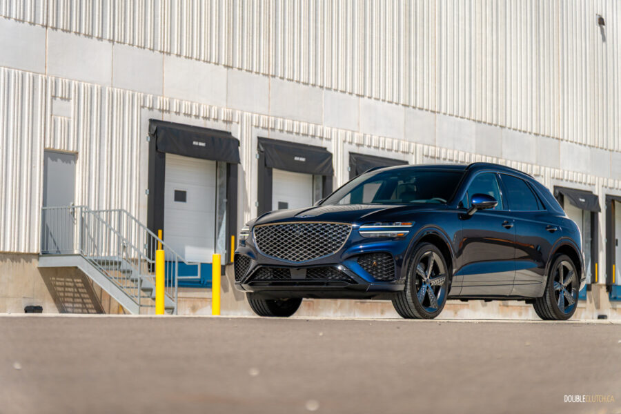Front quarter view of a dark blue 2025 Genesis GV70 3.5T Sport Plus on a sunny day, with a building in the background