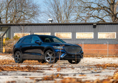 Front quarter view of a dark blue 2025 Genesis GV70 3.5T Sport Plus on a sunny day, with a building in the background