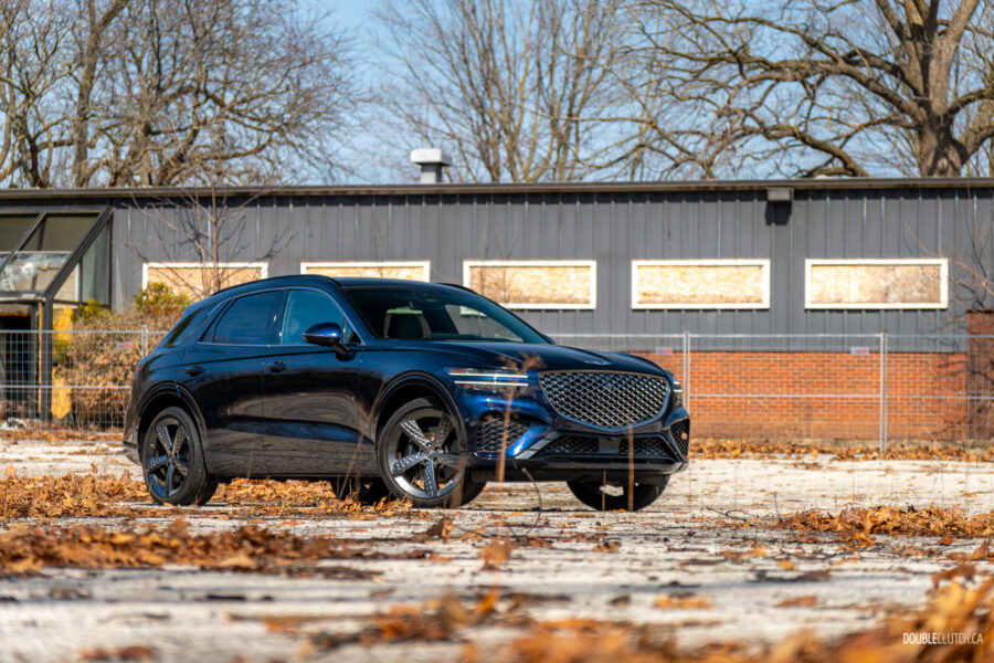 Front quarter view of a dark blue 2025 Genesis GV70 3.5T Sport Plus on a sunny day, with a building in the background