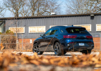 Rear quarter view of a dark blue 2025 Genesis GV70 3.5T Sport Plus on a sunny day, with a building in the background