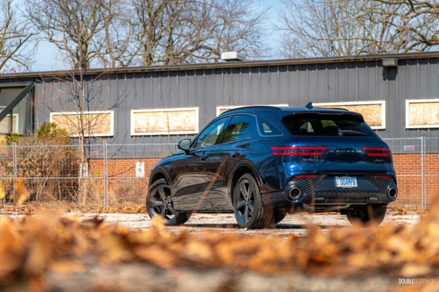 Rear quarter view of a dark blue 2025 Genesis GV70 3.5T Sport Plus on a sunny day, with a building in the background