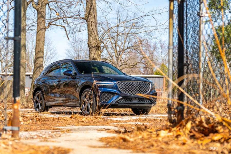 Front quarter view of a dark blue 2025 Genesis GV70 3.5T Sport Plus on a sunny day, with trees in the background