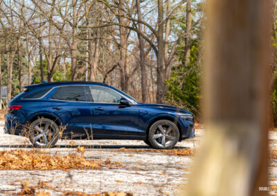Side profile view of a dark blue 2025 Genesis GV70 3.5T Sport Plus on a sunny day, with trees in the background