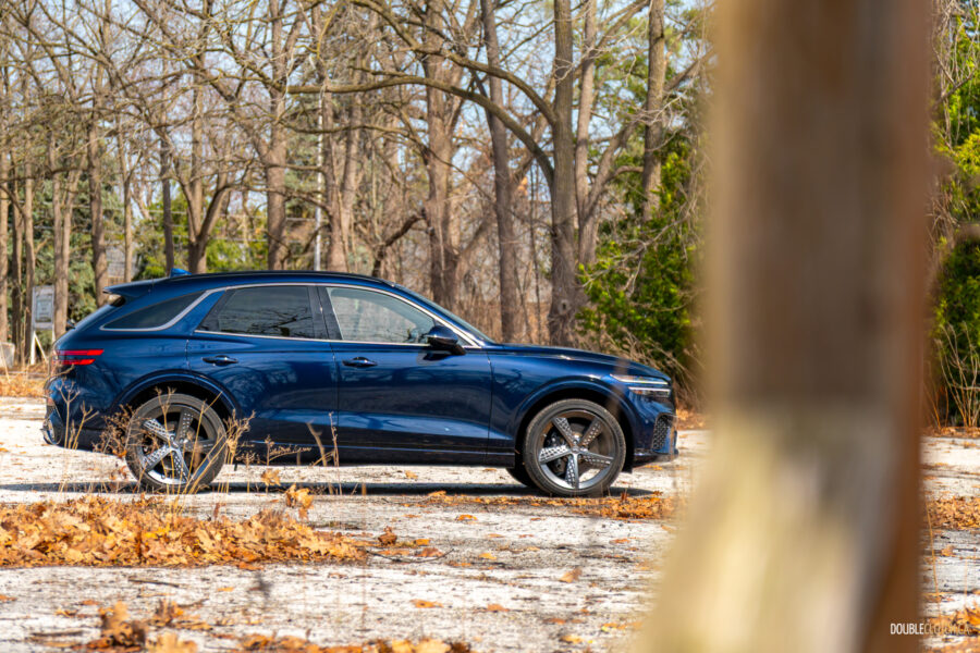 Side profile view of a dark blue 2025 Genesis GV70 3.5T Sport Plus on a sunny day, with trees in the background