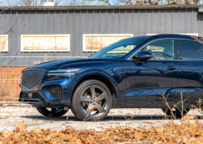 Cropped front quarter view of a dark blue 2025 Genesis GV70 3.5T Sport Plus on a sunny day, with a building in the background