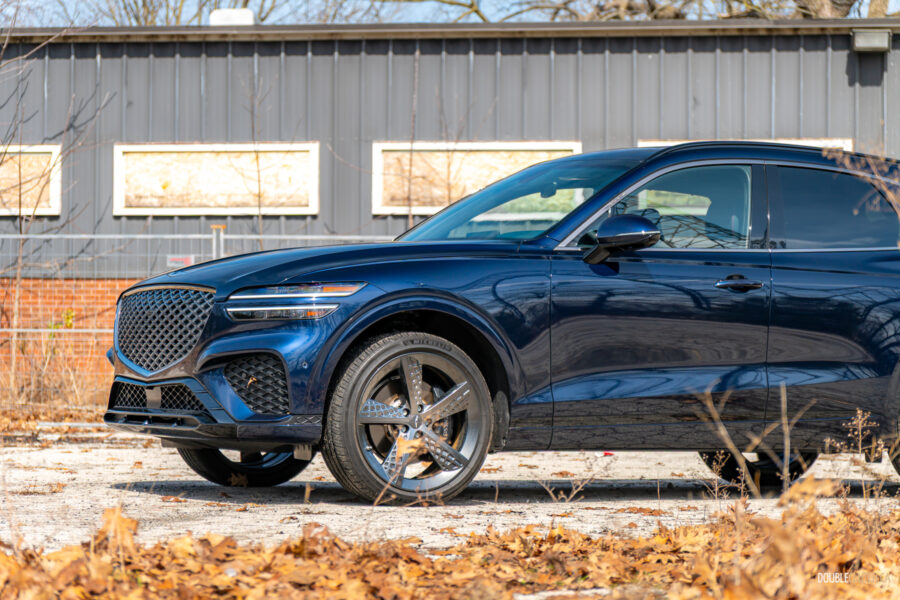 Cropped front quarter view of a dark blue 2025 Genesis GV70 3.5T Sport Plus on a sunny day, with a building in the background