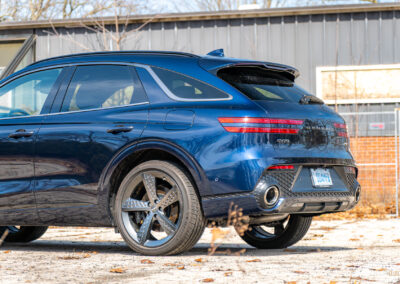 Cropped rear quarter view of a dark blue 2025 Genesis GV70 3.5T Sport Plus on a sunny day, with a building in the background