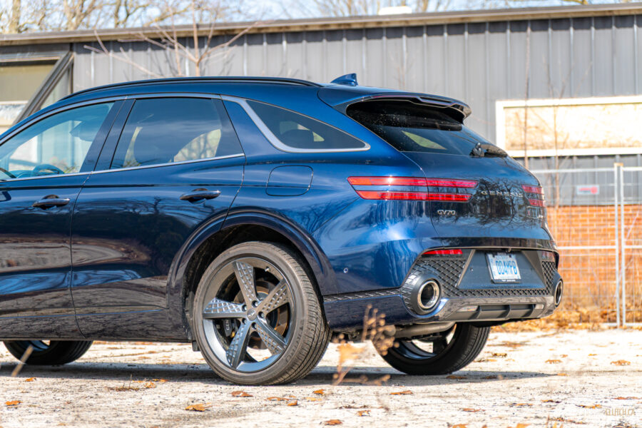 Cropped rear quarter view of a dark blue 2025 Genesis GV70 3.5T Sport Plus on a sunny day, with a building in the background
