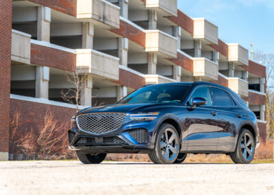 Front quarter view of a dark blue 2025 Genesis GV70 3.5T Sport Plus on a sunny day, with a building in the background